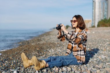 Çakıl taşlı bir sahildeki sıradan kadın fotoğrafçı kamerayla poz veriyor, açık deniz çekimlerinin keyfini çıkarıyor. Sıcak güneş ışığı, kot ve ekose kıyafet, sakin kıyı ortamında uzak yüksek katlı siluetler.