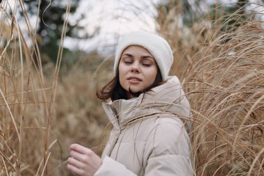 Geniş bir tarlada altın otların ortasında kalın bir palto ve bereli bir kadın, sakin bir poz ve hafif bir esinti doğal tonlarda ve atmosferde sessiz bir açık hava portresini çerçeveliyor..