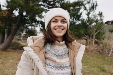 Kışın açık havada gülümseyen kadın, kalın bir palto ve bere giymiş, ağaçlar ve doğal manzarayla çevrili, samimi bir mevsim portresi için..