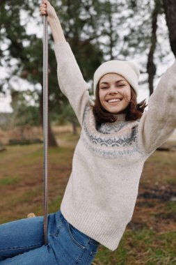Neşeli bir açık hava sahnesi, salıncakta gülümseyen bir kadın, örgü süveter ve beyaz bere, rahat bir ortam, doğal ortam, hafif bir esinti, gençlik ve özgürlük..
