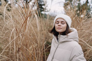Kışlık ceketli ve beyaz bereli bir kadın durgun bir manzarada uzun ve kuru otların arasında duruyor, sakin ve sade bir sadelik yayıyor..