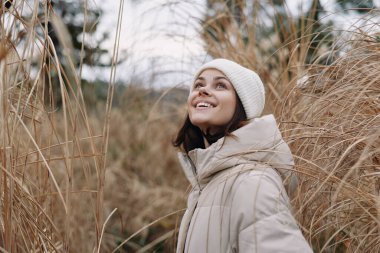 Açık hava portresi, açık renk ceketli ve sazlıklar arasında örgü şapkalı bir kadın, serin mevsimlik ortamda neşeli bir ifade, doğal ve sakin bir ortam.