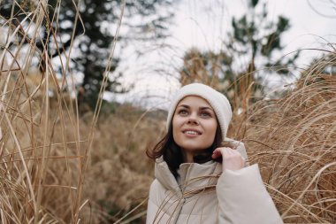 Açık havada, krem rengi ceketli ve bereli bir kadının portresi, uzun otların arasında, soğuk mevsim atmosferi, hafif esinti, barışçıl ifade, doğal dokular..