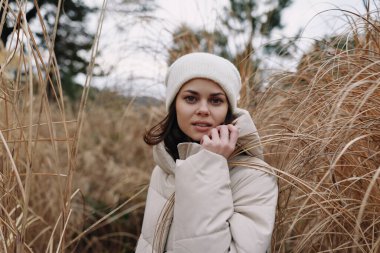 Sade bej ceketli, beyaz örgü şapkalı, uzun otların arasında, sakin, açık hava portresi hafif ve düşünceli mevsimsel atmosferli bir kadının yakın çekimi..