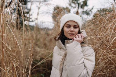 Açık hava sahnesinde kabarık bir ceketi ve şapkası olan bir kadın, sazlıklar arasında telefon görüşmesi, sakin bir ifade ve rüzgarlı bir atmosfer mevsimsel yaşam tarzını işaret ediyor.