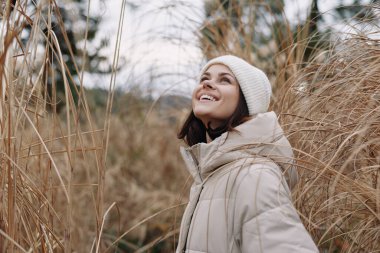 Açık havada kabarık bir ceket ve örgü şapka giyen bir kadının portresi, kış manzarasında uzun kamışların arasında yükselen bir gülümseme ve parlak doğal ışık..