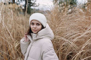 Açık renk ceketli ve bereli bir kadın uzun otların arasında duruyor, açık hava portresi sakin bir havayı, soğuk hava tarzını ve rahat bir zarafeti yansıtıyor.