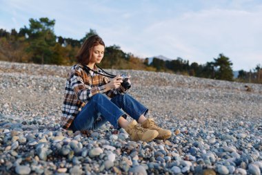 Çakıl taşlı bir sahilde, kameralı, rahat bir poz ve doğal ışık, rüzgarlı saçlar, manzara tasviri, sahil ve huzur dolu bir sahil havası olan bir kadın.