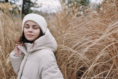 Sıcak bir palto giymiş bir kadının portresi ve uzun çimenler, durgun poz ve doğal ışık arasında örgü bir şapka, açık hava sahnesi dokuyu ve kışa nazik bir sonbahar geçişini vurguluyor..