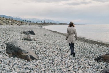 Paltolu ve şapkalı yetişkin bir kadın deniz kıyısında çakıl taşlı sahilde yürüyor ve arka planda bulutlu bir gökyüzü var. Yalnızlık, doğa, seyahat, sakinlik ve açık hava yaşam tarzı kavramı..