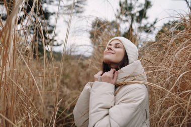 Sıcak bir palto ve örgü eşarplı bir kadının portresi, uzun otların arasında mutlu bir gülümsemeyle duruyor, sakin, sıcak ve doğal bir hava yayıyor.