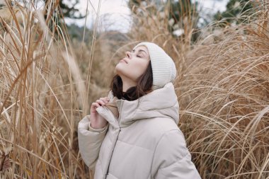 Uzun otlarla çevrili, kalın ceketli ve bereli bir kadının olduğu açık hava sahnesi. Yukarı bakıyor, modayı, doğayı ve sessiz yansıtıcı havayı vurguluyor..