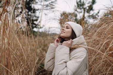Sıcak bir palto içindeki bir kadının portresi ve kışın otların arasında örgü bir bere örmek, sakin bir ruh hali yakalamak, hafif rüzgar ve açık hava modası için nötr tonlar ve doğa ilham verici yaşam tarzı tasvirleri
