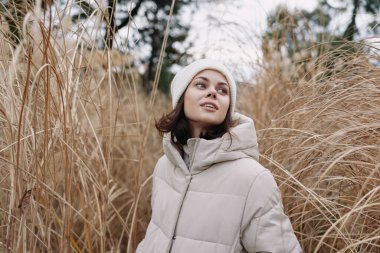 Geniş bir çim tarlasında yastıklı bir palto giyen ve örgü şapkalı bir kadının portresi, dingin bir ifade, hafif bir esinti ve soluk renk havası, açık hava kıyafetleri, seyahat ve ruh hali görüntüleri için ideal..