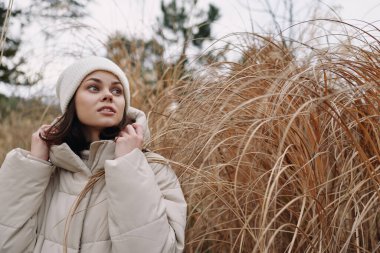 Açık hava sahnesinde, rahat bir dış giyimli genç bir kadın, derin düşüncelere dalmış çimenlerin yanında, kış havasını, hafif esintiyi ve samimi bir portrede sessiz toprak tonlarını yansıtıyor..