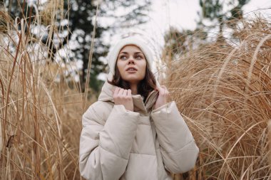 Açık hava sahnesinde, kuru çimenler, durgun poz ve yumuşak ışık arasında sıcak kış ceketi giyen bir kadın moda, doğa ve mevsimsel sükunet için doğal bir zemin yer alıyor.