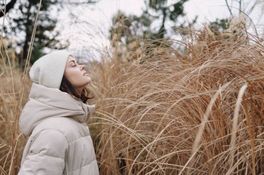 Açık hava kış sahnesinde sıcacık bir ceket içindeki bir kadın seyrek otların arasında, soğuk bir günde doğanın sessiz güzelliğinde dinginlik ve yalnızlığı yakalıyor..