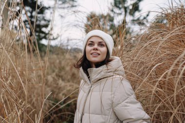 Açık havada bej ceketli ve beyaz bereli bir kadının portresi otların arasında, sakin bir kış havasını, rahat ve doğal atmosferiyle yakalıyor..