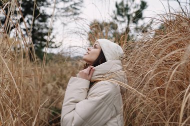 Kuru otlarla çevrili, kalın bir palto ve örgü şapkalı sakin bir kadının olduğu açık hava sahnesi, yumuşak ışığın huzurlu bir ruh hali yaratması gibi yukarı bakıyor..