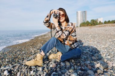 Kayalık bir kıyı şeridindeki kadın fotoğrafçı, deniz manzarası yakalamak için bir kamera kullanıyor, çizmeli ve güneş gözlüklü günlük kıyafetler giymiş, parlak bir gökyüzüne karşı..