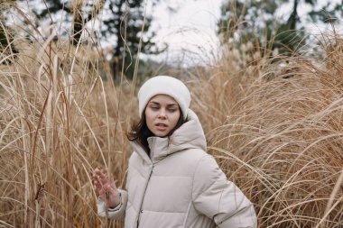 Sıcak kabarık ceketli kız kış boyunca uzun kuru çimenlerin üzerinde duruyor, durgun poz, bulutlu gökyüzü, hafif rüzgar, sessiz tonlar, basit açık hava portresi..