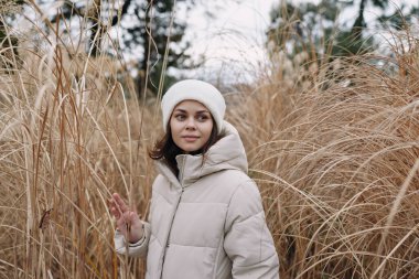 Uzun otların, dingin havanın, kış ortamının, doğa sahnesinin ve hafif esintilerin ortasında, yastıklı bir palto ve örgü şapkalı bir kadının açık hava portresi.