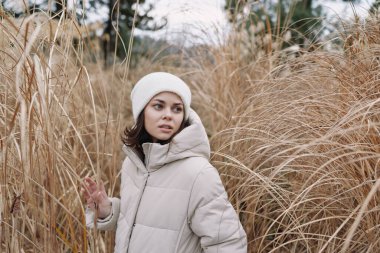 Uzun otların arasında sıcak bir palto giymiş bir kadının sakin açık hava portresi, sonbahar tonlarını, sakin ruh halini ve sakin bir ortamda doğa, yaşam tarzı ve moda temalarını yakalıyor..