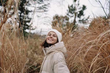 Açık hava kışlık ceketli kadın çimenli bir tarlada gülümsüyor, açık hava modasını ve doğal ortamı vurgulayan samimi bir portre, sakin bir ortamda..