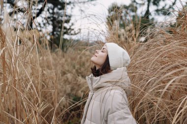 Bej bir ceket giyen bir kadının portresi ve altın otların arasında örgü bir bere, açık doğa ortamında huzurlu bir ruh haliyle yukarı bakıyor.