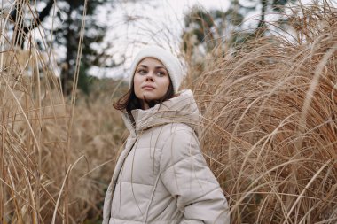 Açık hava sahnesinde kahverengi otların, kış aylarının, dingin havanın ve portrede hazır bekleyen hafif rüzgarın ortasında kabarık ceketli bir kadın görülüyor.
