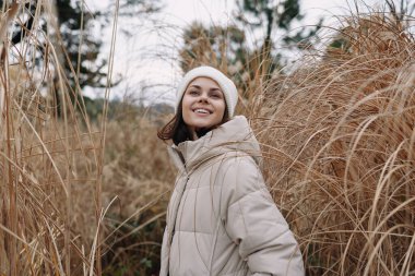 Bej ceketli gülümseyen bir kadının portresi kuru otların arasında duruyor, serin havalarda huzurlu bir açık hava anını yakalıyor..