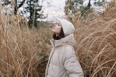 Açık hava kış sahnesinde, uzun otların arasında kalın bir palto giyen ve örgü şapkalı bir kadın, sakin, dingin bir atmosfer ve sessiz tonlarda serin bir ortam sunuyor..