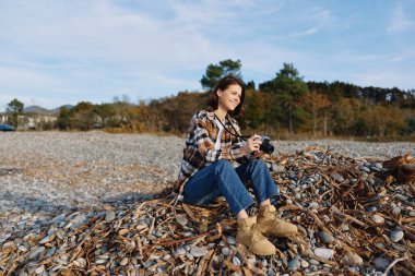 Sahilde yaprak kaplı bir kadın fotoğrafçı, ağaçlarla çevrili el kamerasıyla anları yakalıyor ve sonbaharda açık gökyüzünü seyrediyor.