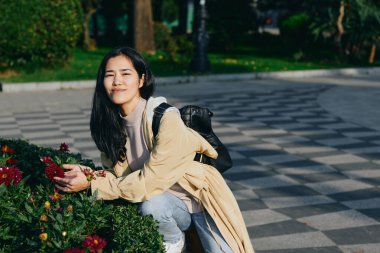 Açık hava parkında çiçeklerin, bej ceketin, sırt çantanın yanında çömelmiş bir kadın, rahat bir gülümseme ve huzurlu bir bahçe havası yaratan yemyeşil bir kadın.