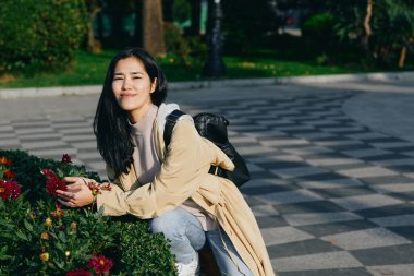 Parkta çiçek açan bir kadınla açık hava sahnesi, gündelik ceket, sırt çantası, güneşli yol, sakin ruh hali, doğa fotoğrafçılığı, samimi an, peyzaj, temiz hava, boş zaman aktivitesi stok görüntüsü,