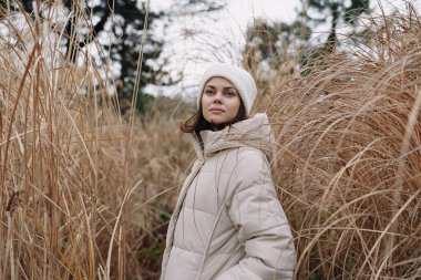 Uzun otların, doğal ışığın, rahat bir duruşun, açık hava modasının ve mevsimsel tarzın ortasında, bej ceketli, huzurlu bir kadının portresi.