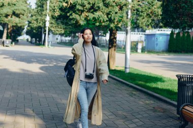 Güneşli bir sokakta günlük gezgin, elinde kamera, uzun ceket ve kot pantolon, arka planda yeşil ağaçlar, gündüz vakti şehir sahnesinde ilerliyor.