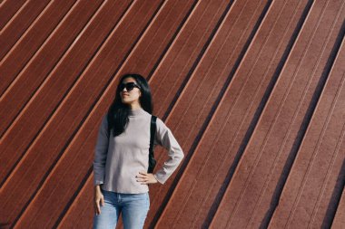 Sıradan giyinmiş bir kadın cesur çapraz bir duvara karşı poz veriyor, güneş gözlükleri takılı, stili, güveni ifade eden, ve güneşli açık hava manzarası ve mimari dokusunda enerjik bir sokak havası olan bir kadın..