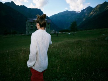 Beyaz örülü bir süveter giymiş bir gezginin arka görüntüsü. Açık bir çayırın ortasında dağlar ve berrak gökyüzü. Doğayı, seyahatleri, yalnızlığı ve kırsal manzarayı çağrıştırıyor.