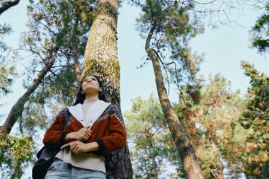 Açık hava orman sahnesi, bir kadının ışığa, şık bir cekete ve eşarba, sakin bir atmosfere, temiz havaya ve orman ortamının sessiz güzelliğine doğru bakan ağaçları gösteriyor.