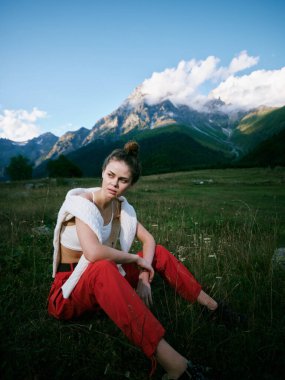 Açık hava sahnesinde, engebeli dağlara karşı çimenlerde, kırmızı pantolonlarda ve beyaz bluzlarda dinlenen bir kadın, modayı, doğayı, seyahatleri ve huzurlu bir manzarada macerayı ön plana çıkarıyor.