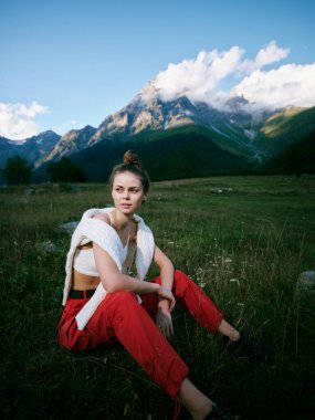 Açık hava sahnesinde, dağ vadisinde çimenlerin üzerinde oturan bir kadın gündelik şık kıyafetler ve doğal manzara manzaranın güzelliğini ve sakin atmosferini ön plana çıkarıyor.