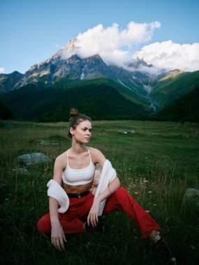 Çimenli, düz ve dağ zeminli açık hava manzarası, beyaz bluz ve kırmızı pantolonla poz veren ileri moda kadın, sakin ruh hali, yumuşak ışık, temiz hava, vahşi doğa ve ilham verici stil.