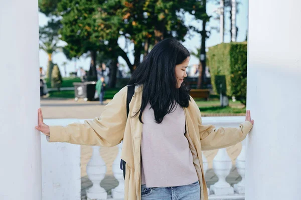 Açık hava sahnesinde, mimari sütunlar arasında bej bir palto giyen, binaların üzerinde dinlenen eller, uzağa bakan, yeşil ve yumuşak güneş ışığı olan sakin park atmosferi olan bir kadın görülüyor.