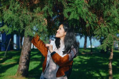 Ağaçlar ve yeşillikler arasında gülümseyen bir kadının yer aldığı açık hava sahnesi, sallanan yapraklar, sıcak güneş ışığı ve sakin atmosfer ile etkileşim, rahatlama, neşe ve doğa ile uyum göstermektedir.