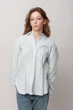 Beyaz gömlekli ve kot pantolonlu genç bir kadının portresi parlak arka planda izole edilmiş, büzüşmüş dudaklarıyla neşeli bir yüz yapıyor. Sıradan moda ve eğlenceli ifade konsepti.