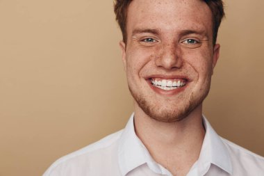 Kızıl saçlı ve çilli mutlu genç adam beyaz gömlekli geniş bir şekilde gülümsüyor. Bej arka planda, doğal ışıklandırmayla yakın plan portresi. Güven, gençlik ve olumlu duygular kavramı.