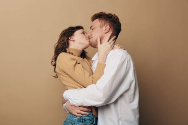 Sıradan giyinen çiftler, tarafsız arka planda nazik bir öpücük ve kucaklaşmayı paylaşırlar. Romantik aşk, samimiyet ve duygusal bağ sıcak, samimi bir atmosferde yakalanır..