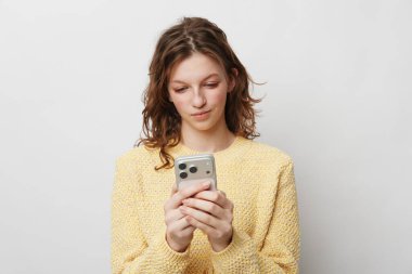 Sarı örgü süveter giyen kadın akıllı telefonu iki eliyle tutup ekrana odaklıyor. Basit arka plan modern iletişimi, dijital cihaz kullanımını ve günlük yaşamı vurgular.