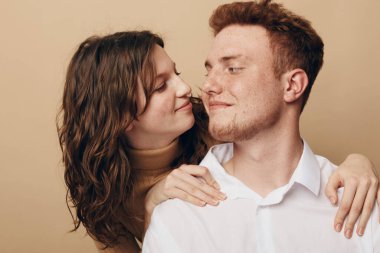 Genç bir çiftin yakın plan fotoğrafı, dalgalı saçlı bir kadın kısa saçlı bir adama nazikçe sarılıyor. Bej arka planda sıcak, sevgi dolu bir etkileşim, romantizm ve duygusal bağ kavramı.